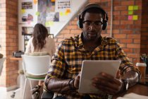 Vista frontal del empresario afroamericano usando tableta digital con auriculares en la oficina mientras una mujer caucásica trabaja detrás de él - foto de stock