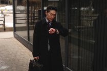 Вид спереди молодого хорошо одетого азиатского бизнесмена, смотрящего на свой смартфон во время прогулки по улице в городе — стоковое фото