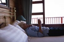 Seitenansicht einer traurigen aktiven Seniorin, die ihr Mobiltelefon benutzt, während sie zu Hause im Schlafzimmer auf dem Bett liegt — Stockfoto