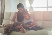 Vista frontal de madre de raza mixta usando hijab e hija usando tableta digital en la sala de estar en casa - foto de stock