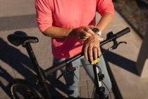 Mittelteil einer aktiven Seniorin, die ihre Smartwatch benutzt, während sie ihr Fahrrad auf einem Spazierweg in der Sonne hält — Stockfoto