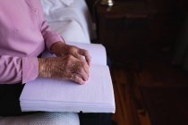 Sección media de una anciana ciega activa leyendo un libro braille con los dedos mientras está sentada en su cama en el dormitorio de su casa - foto de stock