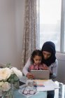 Vista pensativa frontal da mãe de raça mista vestindo hijab e filha usando tablet digital em casa sentado em torno de uma mesa — Fotografia de Stock