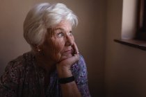 Vorderansicht einer nachdenklichen, aktiven Seniorin, die durch das Fenster blickt und ihr Kinn an ihre Hand in der heimischen Küche lehnt — Stockfoto