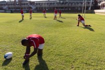 Vista frontale del giocatore di rugby afroamericano che lega i lacci delle scarpe sul campo da rugby con i membri della squadra sullo sfondo nella giornata di sole — Foto stock