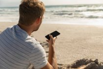 Вид сзади расслабленного мужчины, сидящего на пляже во время разговора по мобильному телефону — стоковое фото