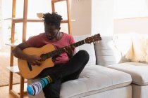 Frontansicht eines afrikanisch-amerikanischen Mannes, der Gitarre spielt, während er zu Hause im Wohnzimmer sitzt — Stockfoto