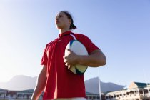 Vue en angle bas d'un joueur de rugby caucasien tenant une balle de rugby et se tenant debout dans le stade par une journée ensoleillée — Photo de stock