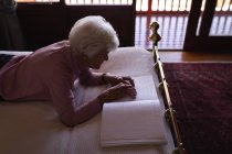 Vista de ángulo alto de una mujer mayor ciega activa acostada en la cama y leyendo un libro braille con los dedos en el dormitorio en casa - foto de stock