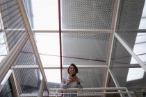 Vista a basso angolo di una donna d'affari asiatica che utilizza il suo telefono cellulare e si appoggia alle barriere di sicurezza in ufficio — Foto stock