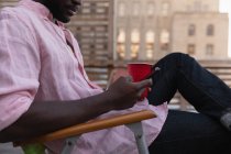 Seitenansicht eines afrikanisch-amerikanischen Mannes, der sein Mobiltelefon benutzt, während er zu Hause auf dem Balkon ein kaltes Getränk trinkt — Stockfoto