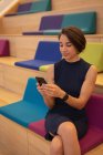 Vista frontal de uma bela mulher de negócios asiática usando seu telefone celular enquanto sentada em escadas no escritório — Fotografia de Stock