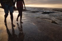 Sección baja de pareja afroamericana caminando sobre sable y relajándose junto al mar. De la mano - foto de stock