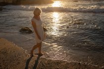 Seitenansicht einer aktiven Seniorin, die am Abend am Ufer des Strandes spazieren geht, mit einem Sonnenuntergang, der sich im Wasser spiegelt — Stockfoto