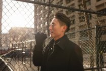 Vue latérale du jeune homme d'affaires asiatique parlant sur un téléphone portable tout en passant devant une clôture à mailles de chaîne dans la rue de la ville — Photo de stock