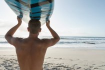 Vista traseira do jovem surfista carregando prancha de surf na praia em um dia ensolarado. Ele está olhando as ondas — Fotografia de Stock