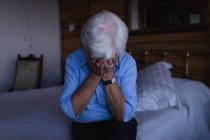 Vorderansicht einer traurigen aktiven Seniorin, die ihr Gesicht mit den Händen bedeckt, während sie zu Hause auf dem Bett im Schlafzimmer sitzt — Stockfoto