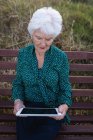 Вид спереду активної старшої жінки, використовуючи цифровий планшет, сидячи на дерев'яній лавці на пляжі — стокове фото