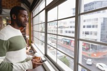 Vue latérale d'un homme d'affaires afro-américain réfléchi utilisant un téléphone portable et tenant un café dans une cantine de bureau — Photo de stock