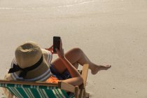 Vista de alto ângulo da jovem mulher relaxando na espreguiçadeira na praia em um dia ensolarado. Ela está sentada e usando seu telefone celular — Fotografia de Stock