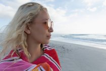 Vista lateral de la mujer rubia de pie en la playa en un día soleado. Lleva gafas solares y observa el océano. - foto de stock