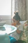 Primer plano de la chica de raza mixta comer fruta naranja sentado en la silla en casa - foto de stock