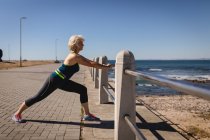 Side view of an active senior woman stretching against the guard rail of the promenade along the beach under the sunshine — Stock Photo