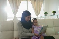 Vue de face de mère mixte portant hijab et fille utilisant une tablette numérique sur le canapé à la maison — Photo de stock