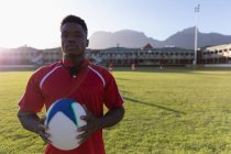 Frontansicht eines afrikanisch-amerikanischen Rugby-Spielers, der den Rugby-Ball in der Hand hält und an einem sonnigen Tag im Stadion steht — Stockfoto