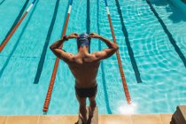 Hochwinkelaufnahme kaukasischer Schwimmer, der auf dem Startblock steht und im Schwimmbad bei Sonnenschein eine Schwimmbrille trägt — Stockfoto
