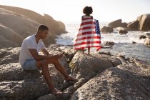 Visão traseira do casal afro-americano relaxando na praia em um dia ensolarado com a menina segurando bandeira americana — Fotografia de Stock