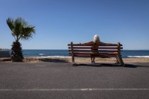 Rückansicht einer aktiven Seniorin, die es sich auf einer Bank in Strandnähe unter der Sonne gemütlich macht — Stockfoto