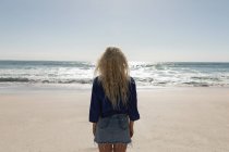Вид сзади на красивую блондинку, стоящую на пляже в солнечный день. Она смотрит на океан. — стоковое фото