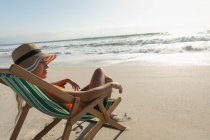 Вид сбоку расслабленной молодой женщины, отдыхающей на солнцепеке на пляже в солнечный день. Она смотрит в пейзаж — стоковое фото