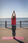 Rückansicht einer aktiven Seniorin, die Yoga auf einer Fitnesskarte auf einer Promenade am Meer macht — Stockfoto