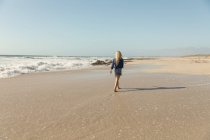 Visão traseira da mulher loira relaxada andando na praia em um dia ensolarado — Fotografia de Stock