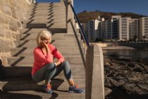 Vorderansicht einer nachdenklichen aktiven Seniorin, die es sich auf der Treppe nahe der Promenade gemütlich macht und in der Sonne aufs Meer blickt — Stockfoto