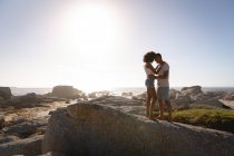 Vista lateral de la pareja afroamericana en un ambiente romántico de pie en la roca cerca del mar. Están cara a cara y mirándose. - foto de stock