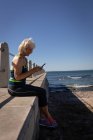 Вид сбоку активной пожилой женщины, которая пользуется мобильным телефоном и веселится, сидя на краю набережной под солнцем — стоковое фото