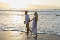 Vista lateral de la joven pareja de amor cogida de la mano mientras está de pie en la playa en un día soleado. Caminan junto al mar, mano a mano - foto de stock