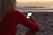 Вид сзади на блондинку с мобильного телефона на пляже. Она сидит на песке. — стоковое фото