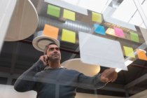 Niedrigwinkel-Ansicht eines Geschäftsmannes, der auf seinem Mobiltelefon spricht, während er auf klebrige Zettel blickt, die an der Wand des Büros befestigt sind — Stockfoto