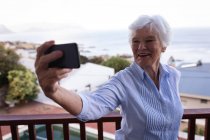 Вид спереди счастливой активной пожилой женщины, делающей селфи на балконе на фоне морского пейзажа со своим мобильным телефоном дома — стоковое фото