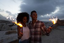 Vista frontal do casal afro-americano curtindo e sorrindo com fogueira na praia após o pôr do sol — Fotografia de Stock