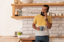 Front view of mixed race Man talking on mobile phone while standing at home in kitchen room — Stock Photo