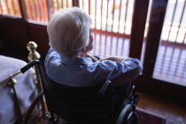Vista posteriore di una donna anziana disabile attiva seduta su una sedia a rotelle e che guarda attraverso la finestra in camera da letto a casa — Foto stock