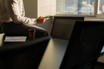 Средняя секция мужчины исполнительной работы на мобильный телефон, сидя на столе и наслаждаясь чашкой кофе в современном офисе — стоковое фото