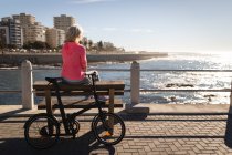 Rear view of an active senior woman relaxing on a bench and looking at the sea on promenade under the sunshine — Stock Photo