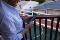 Средняя часть активной пожилой женщины, использующей свой мобильный телефон, стоя на балконе дома — стоковое фото