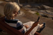 Высокий угол обзора активной пожилой женщины с помощью мобильного телефона, сидящей на скамейке на набережной вечером — стоковое фото